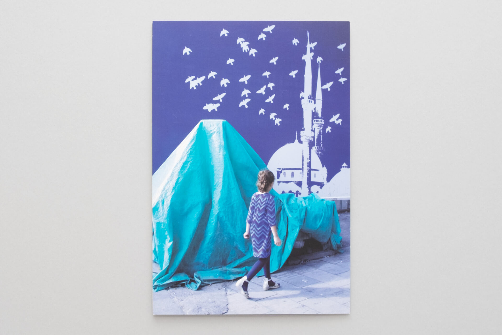 Ausstellungskarte mit Foto aus Istanbul für Floating Volumes: Mädchen läuft vor verhülltem Objekt und schematischem Motiv mit Moschee und Tauben.